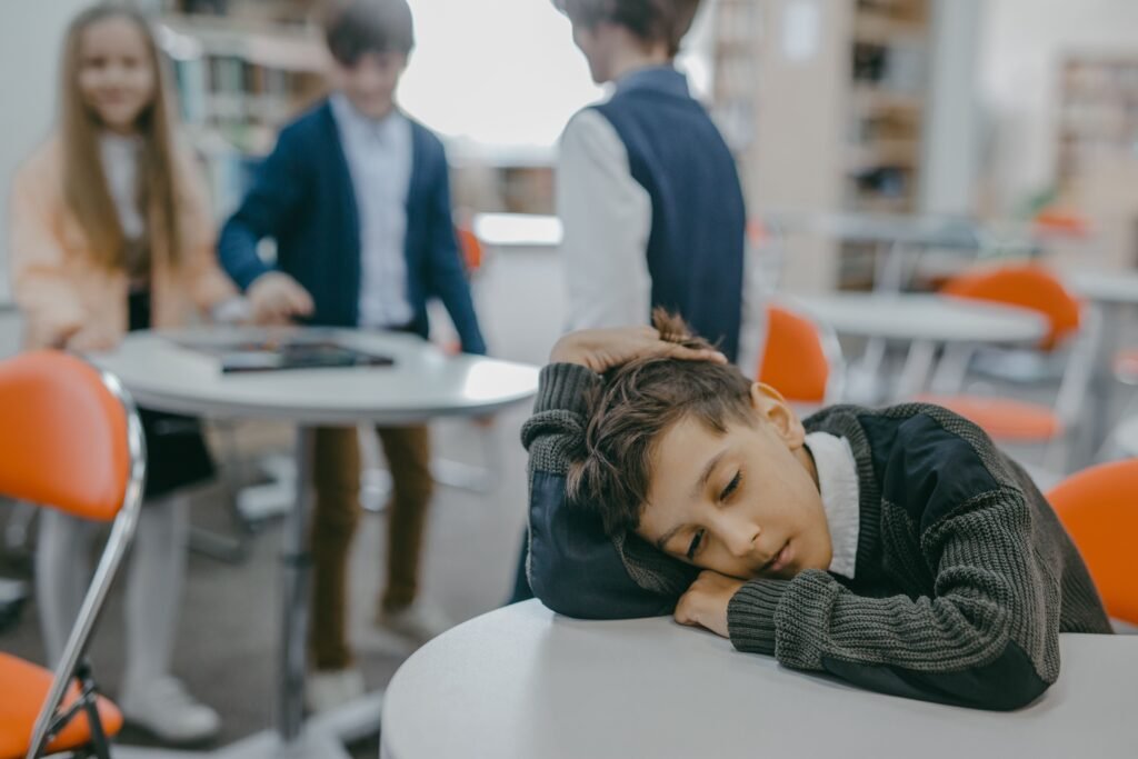 A sad child rests on a classroom desk while peers talk, symbolizing loneliness and school exclusion.